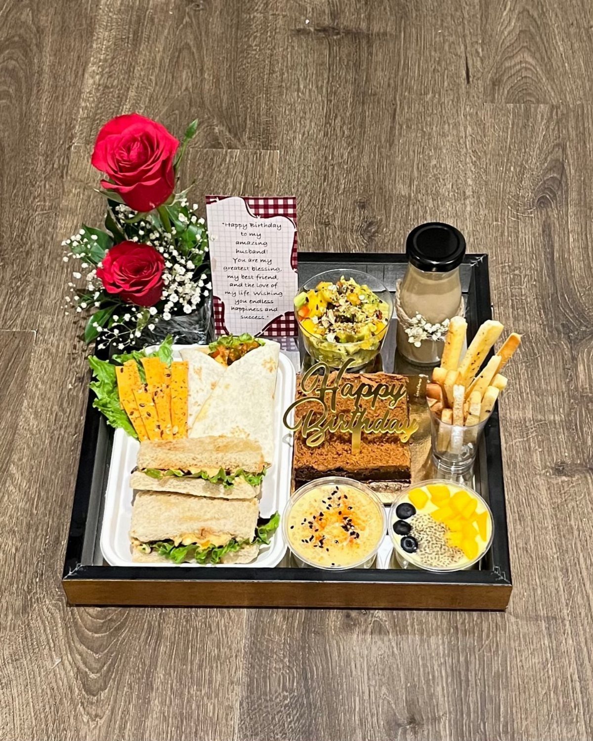 Breakfast tray with sandwiches, cake, fruit, and red roses on a wooden floor