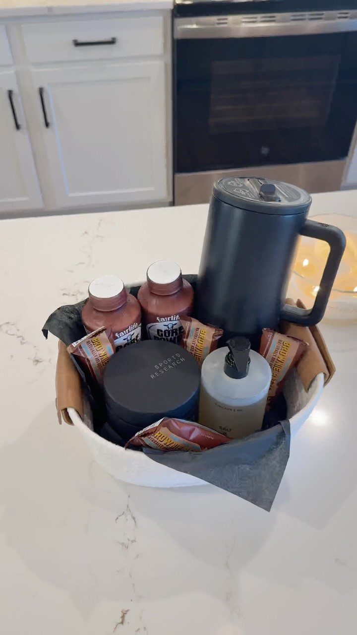 White gift basket containing a French press, coffee bottles, and snacks on marble.