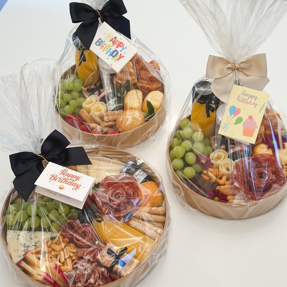 Three birthday charcuterie baskets with meats, cheeses, grapes, and juice wrapped in cellophane.