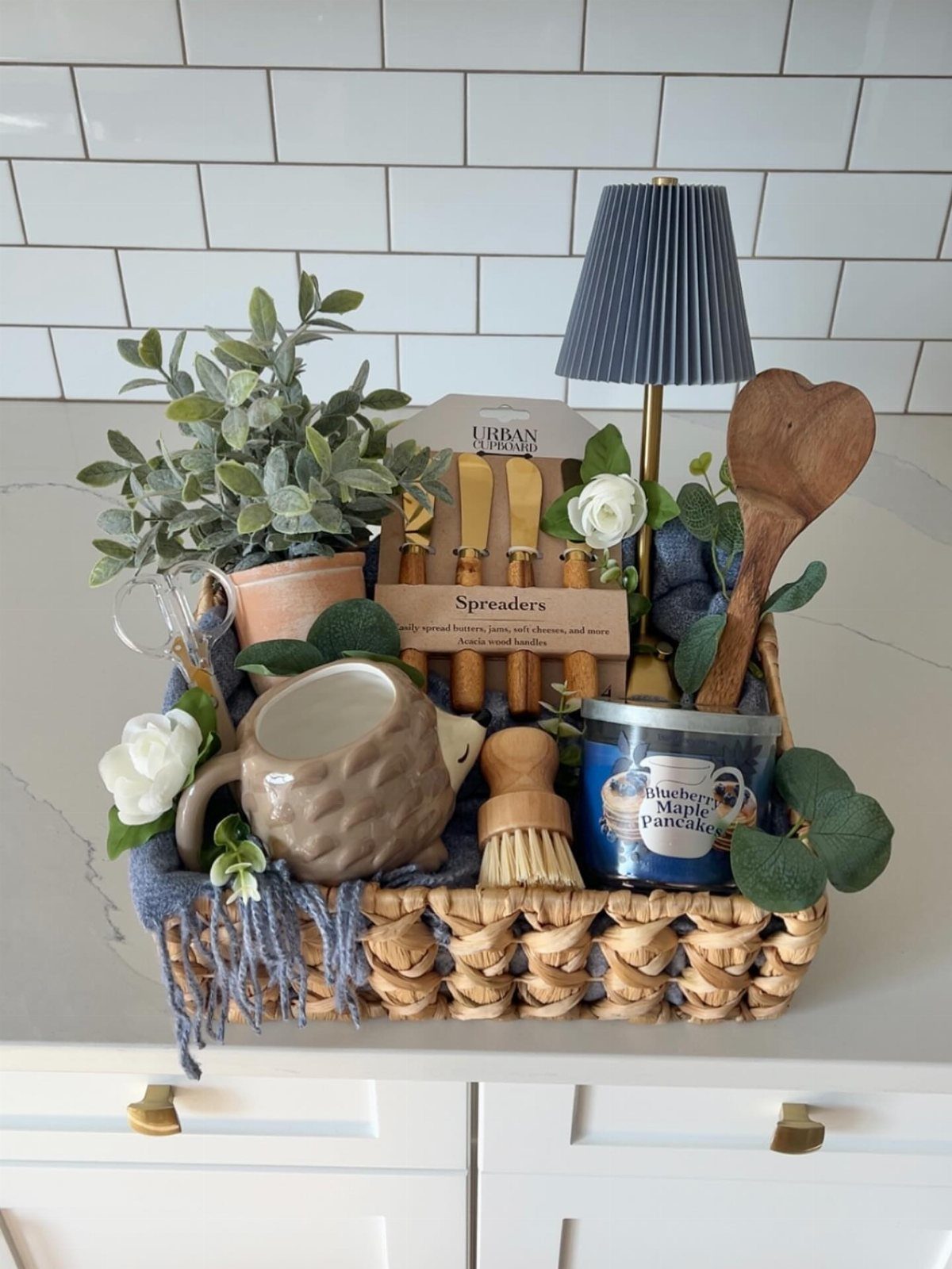 Woven gift basket containing a hedgehog pitcher, plant, lamp, and kitchen accessories.