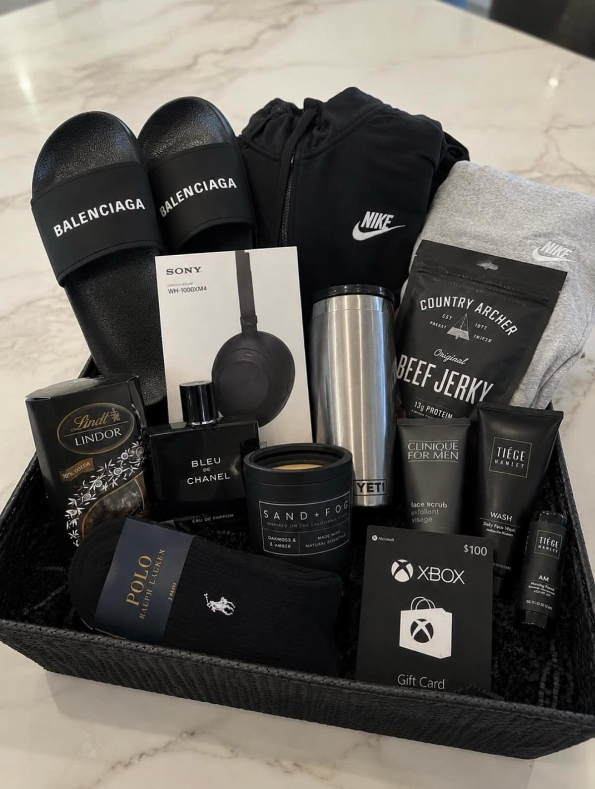 Black gift basket containing luxury clothing, electronics, grooming products, and snacks on marble.