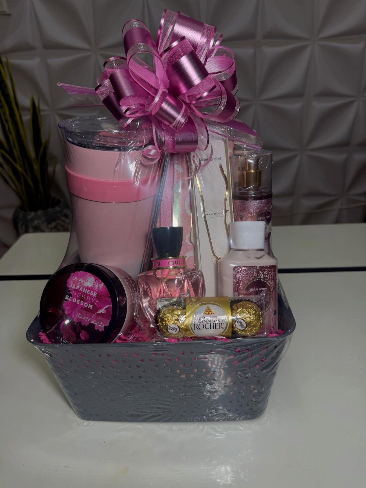 Silver gift basket with pink tumbler, perfume, and chocolates topped with a pink bow.