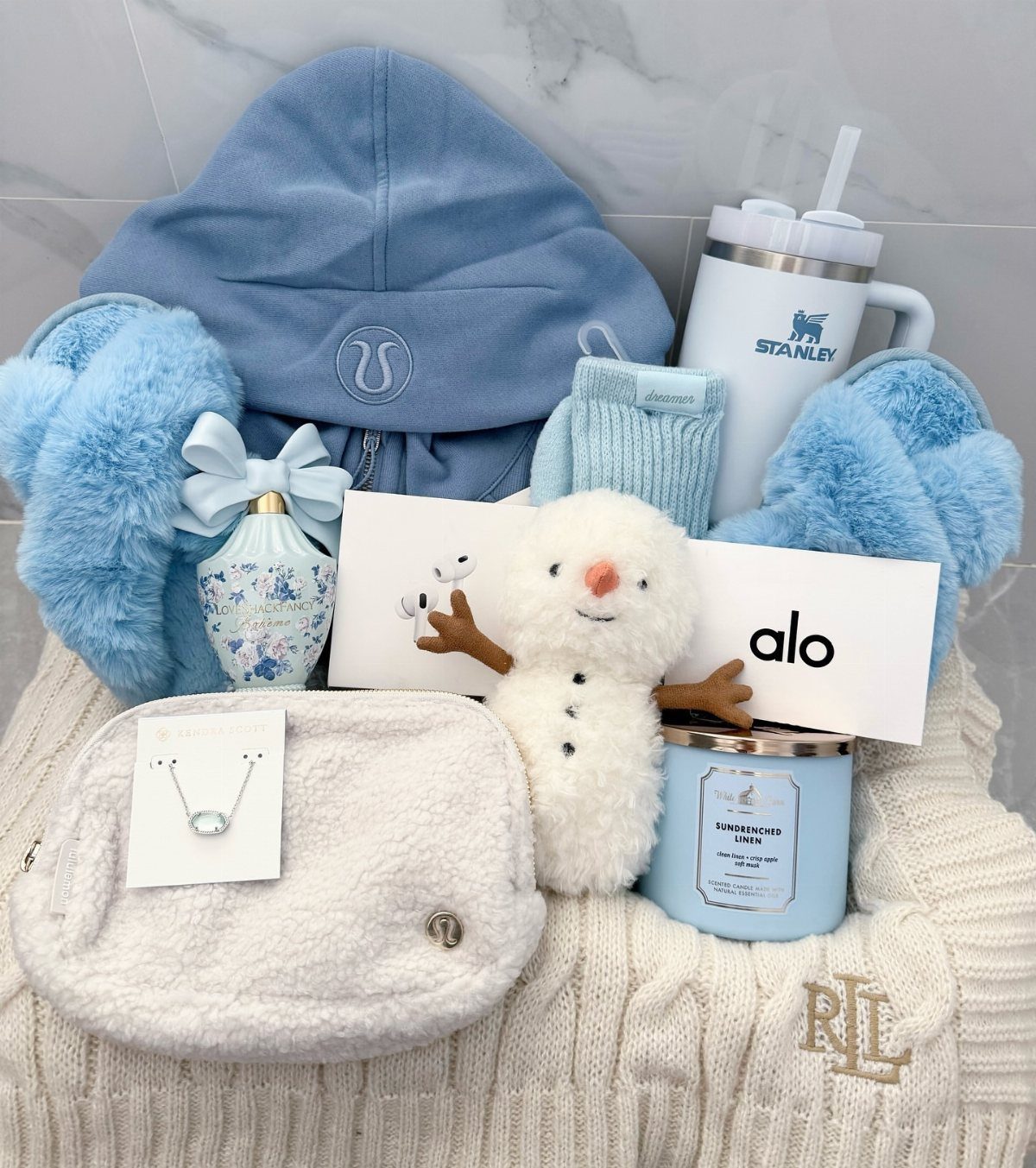 Gift basket with blue fuzzy slippers, plush snowman, and blue hoodie on white sweater.
