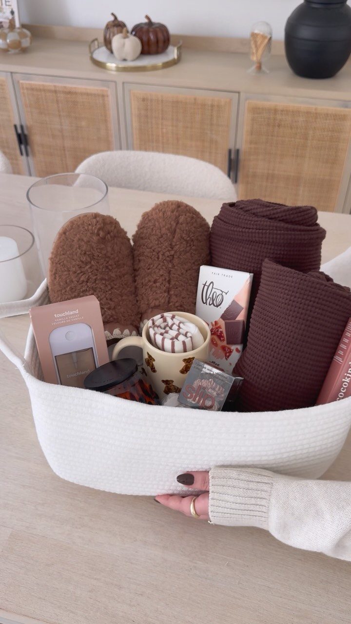 White woven gift basket containing brown fuzzy slippers, blankets, and hot chocolate treats.