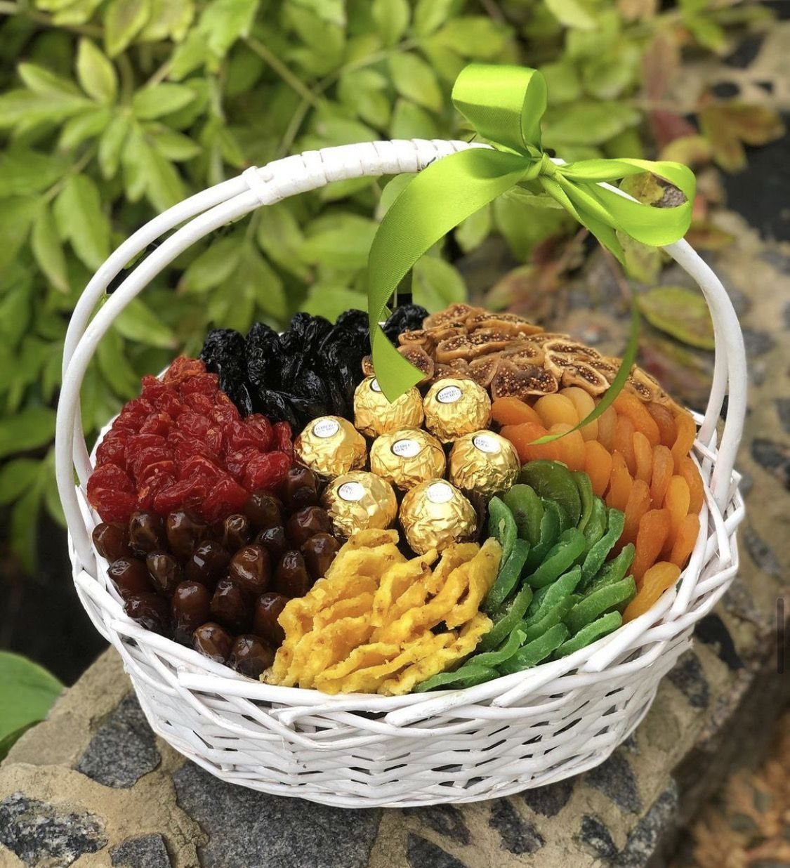 White wicker basket filled with dried fruits and gold-wrapped chocolates with green ribbon.