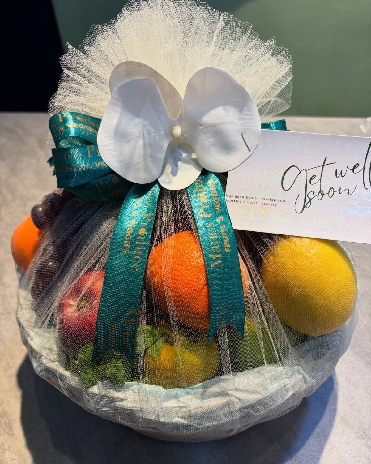 Fruit gift basket wrapped in white tulle with a teal ribbon and get well card.