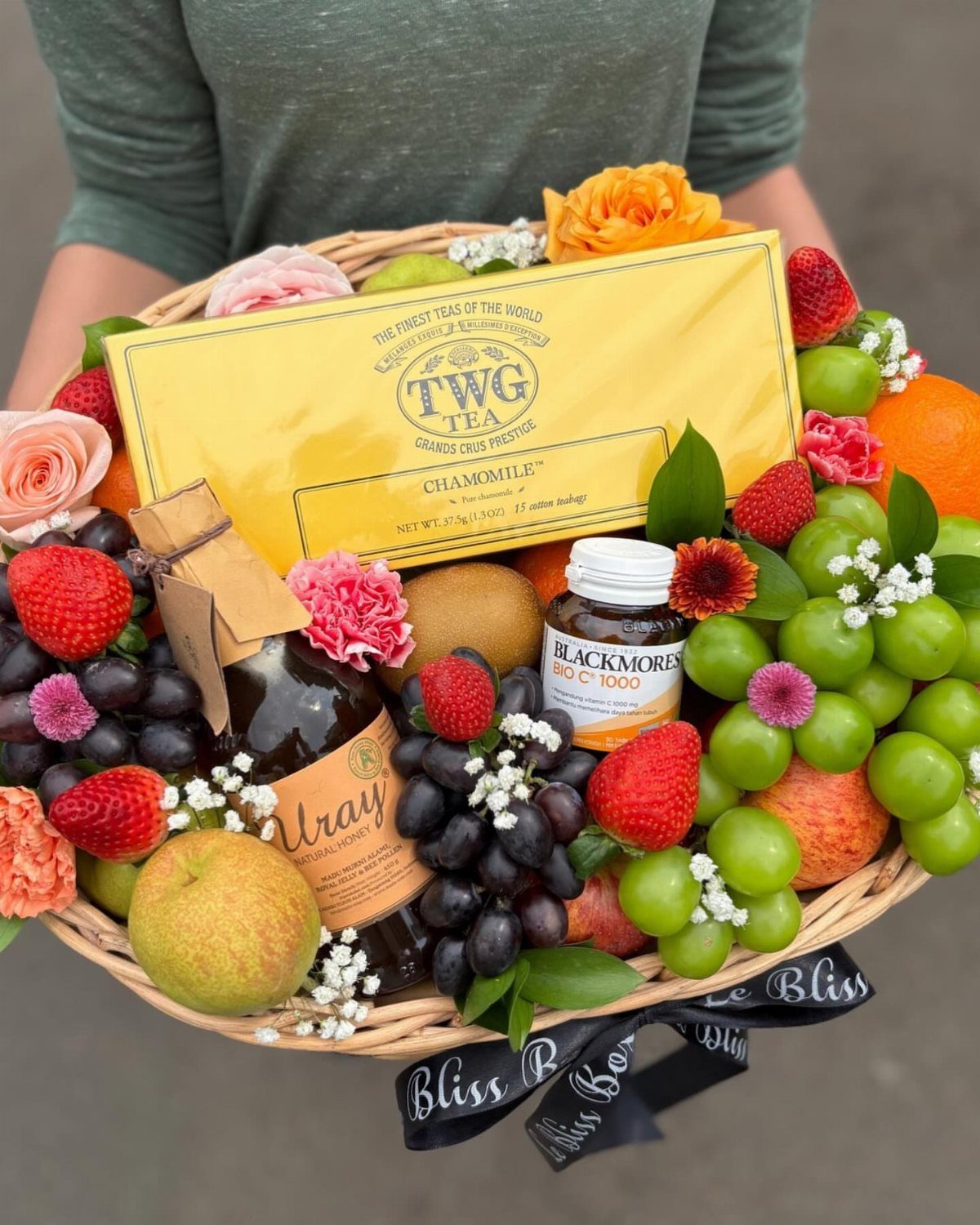 Wicker gift basket containing chamomile tea, vitamins, fresh fruits, and orange roses.