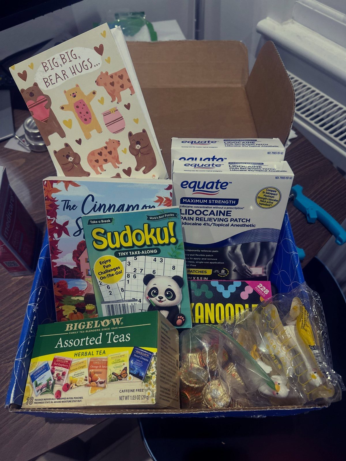 Care package box containing tea, puzzle books, a greeting card, and pain patches.
