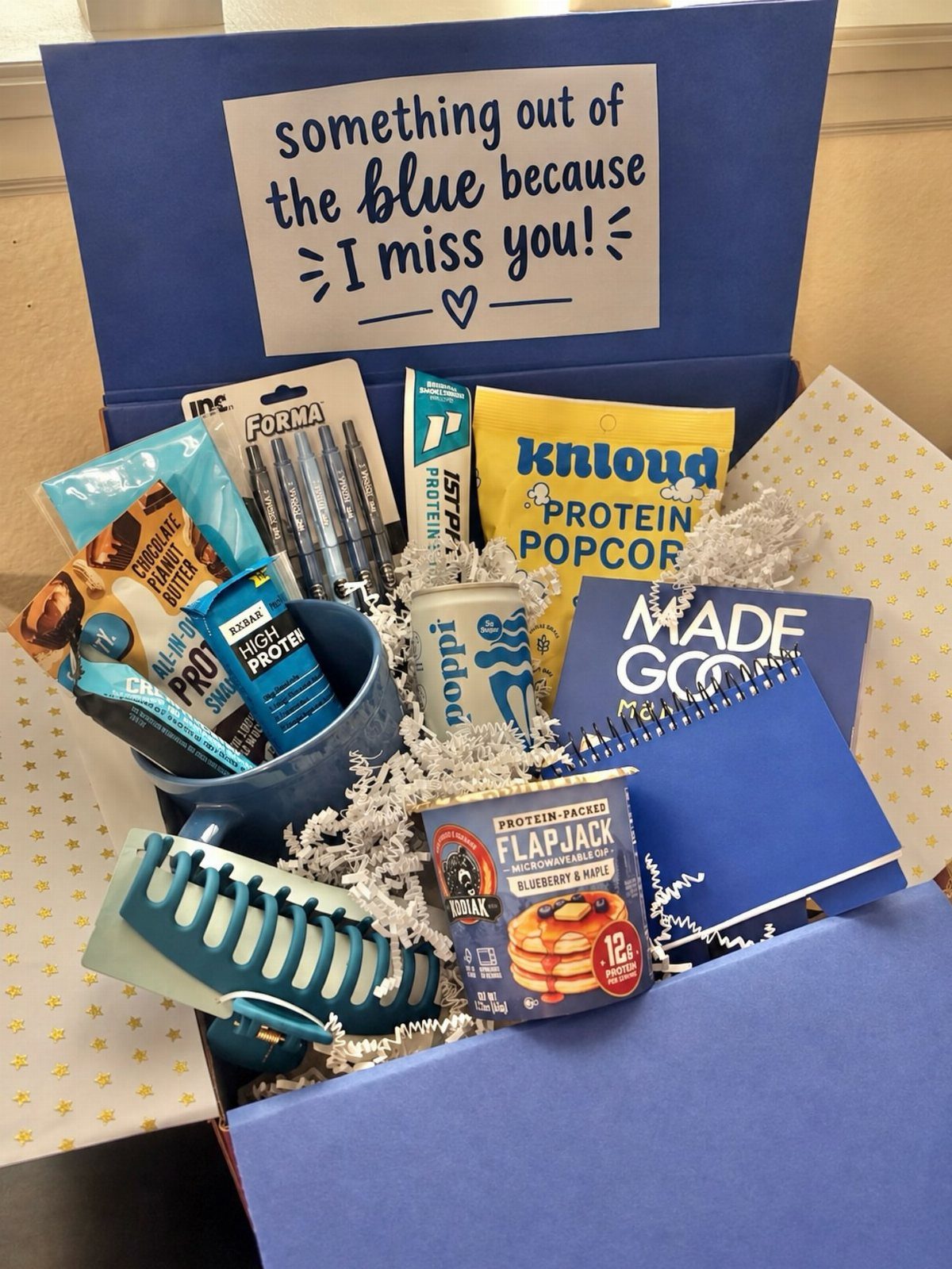 Blue care package box filled with blue snacks, a notebook, and a mug.