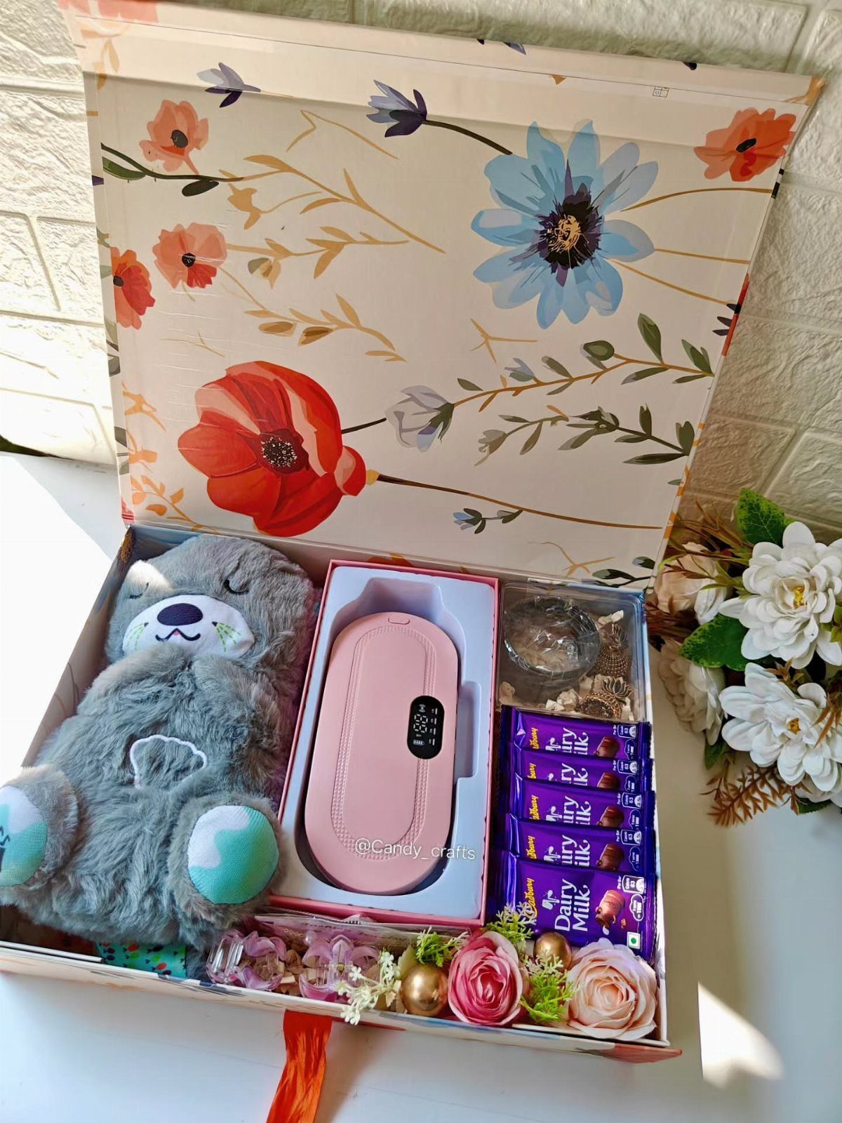 Gift box with a grey teddy bear, pink gadget, chocolates, and artificial flowers.