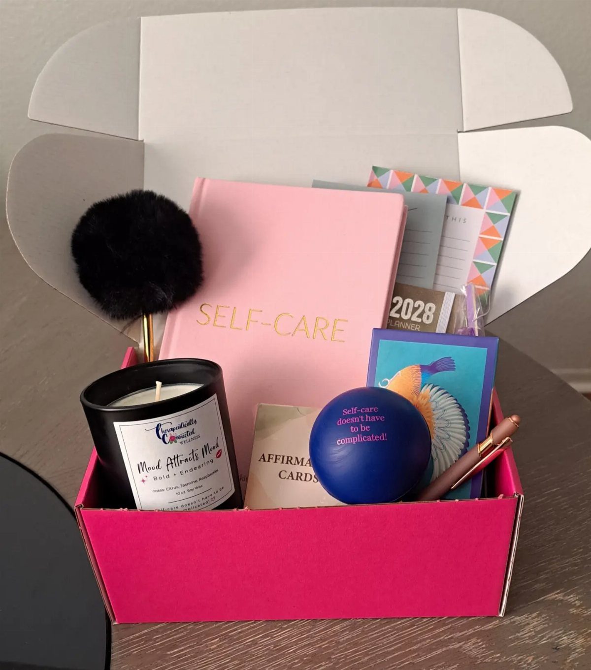 Self-care gift box with pink journal, black candle, blue stress ball, and affirmation cards.