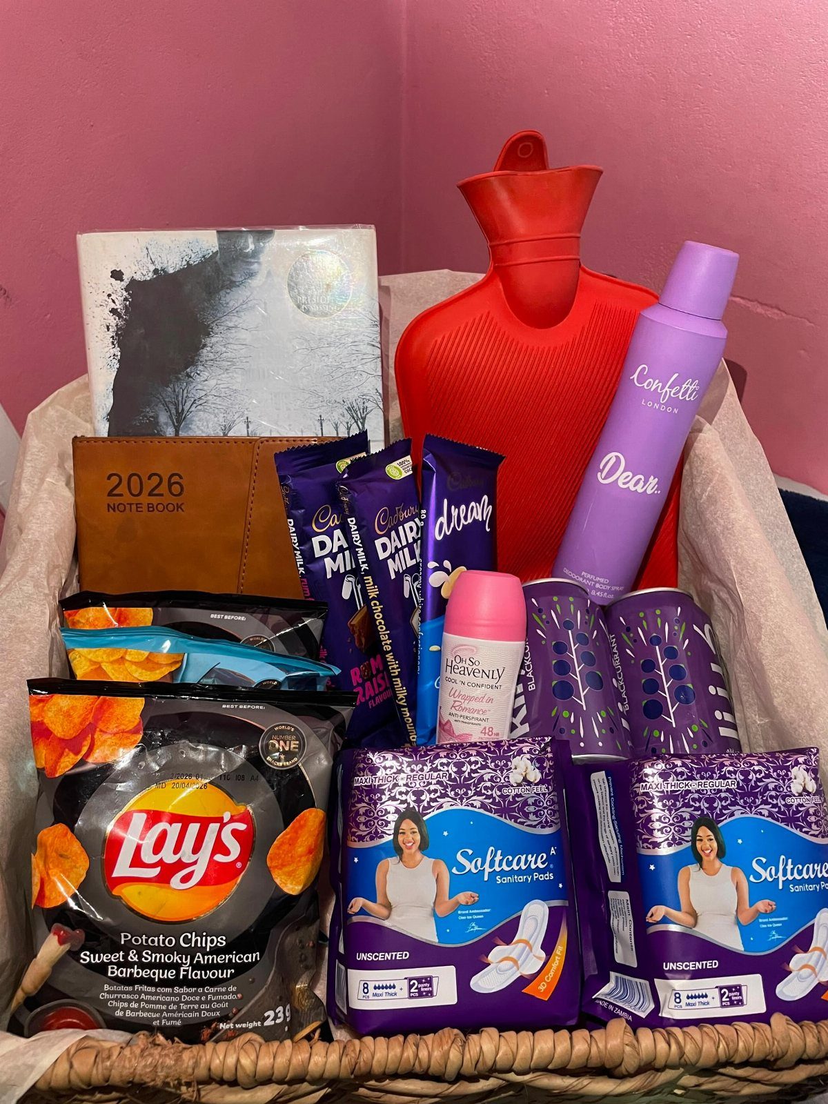 Wicker gift basket containing a red hot water bottle, snacks, and hygiene products.