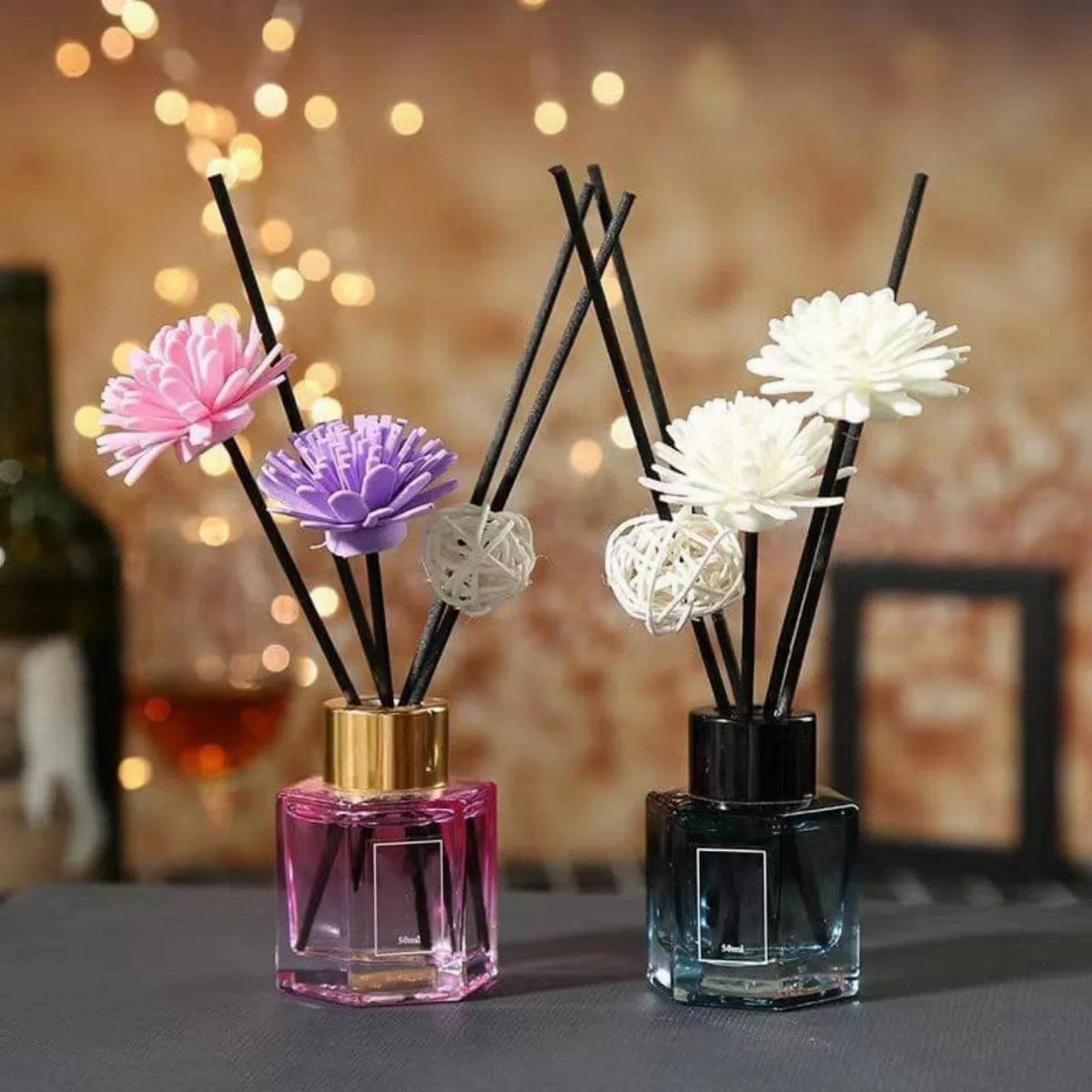 Two reed diffuser bottles with decorative flowers and black sticks on a grey surface.