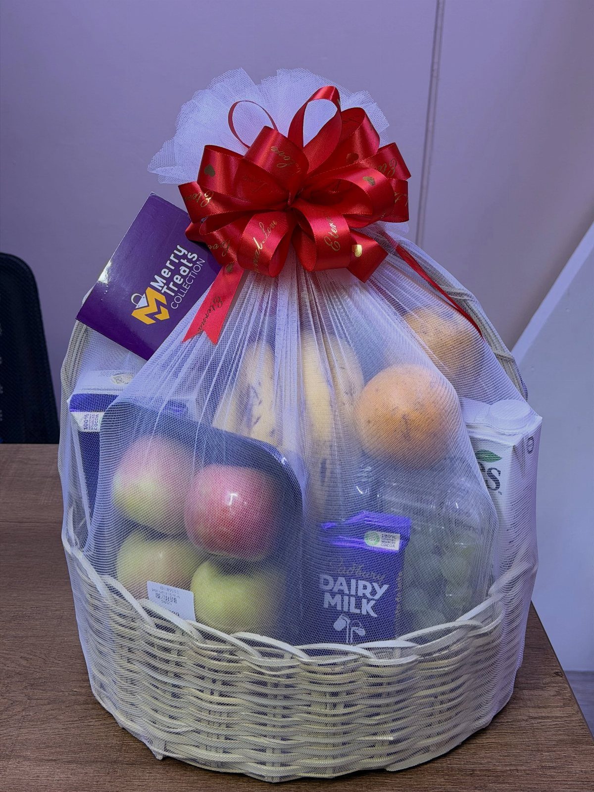 Fruit basket with apples and oranges wrapped in white mesh with red bow.
