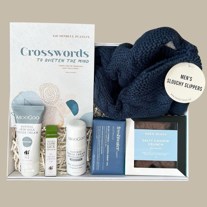 Gift box with crossword puzzle book, navy slippers, skincare products, and cashew snacks.