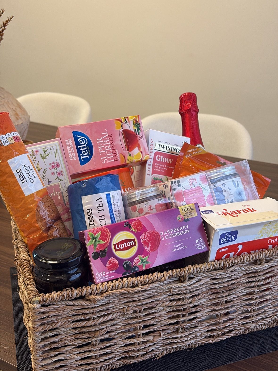Woven wicker gift basket filled with assorted tea, snacks, and a red bottle.