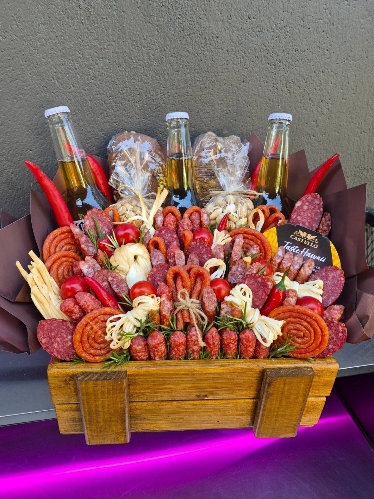 Wooden crate filled with assorted cured meats, sausages, cherry tomatoes, and bottled beer.