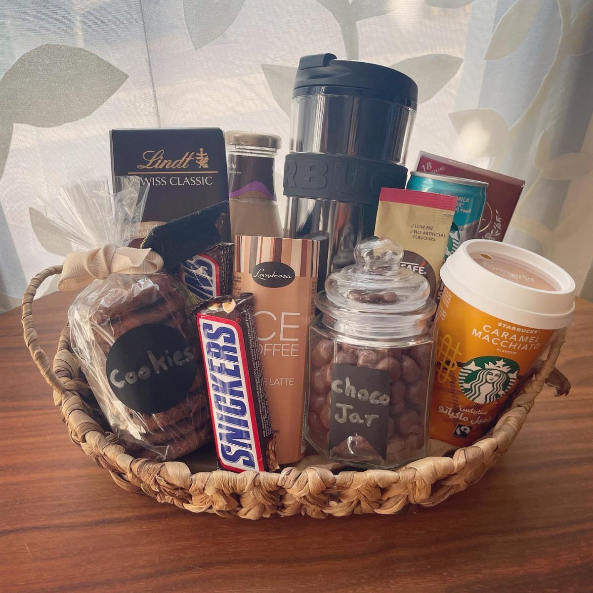 Woven gift basket containing coffee drinks, a travel mug, and various chocolate candies.