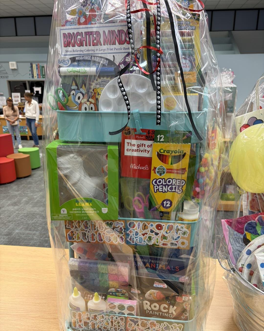 Children's craft gift basket wrapped in plastic containing colored pencils and painting kits.
