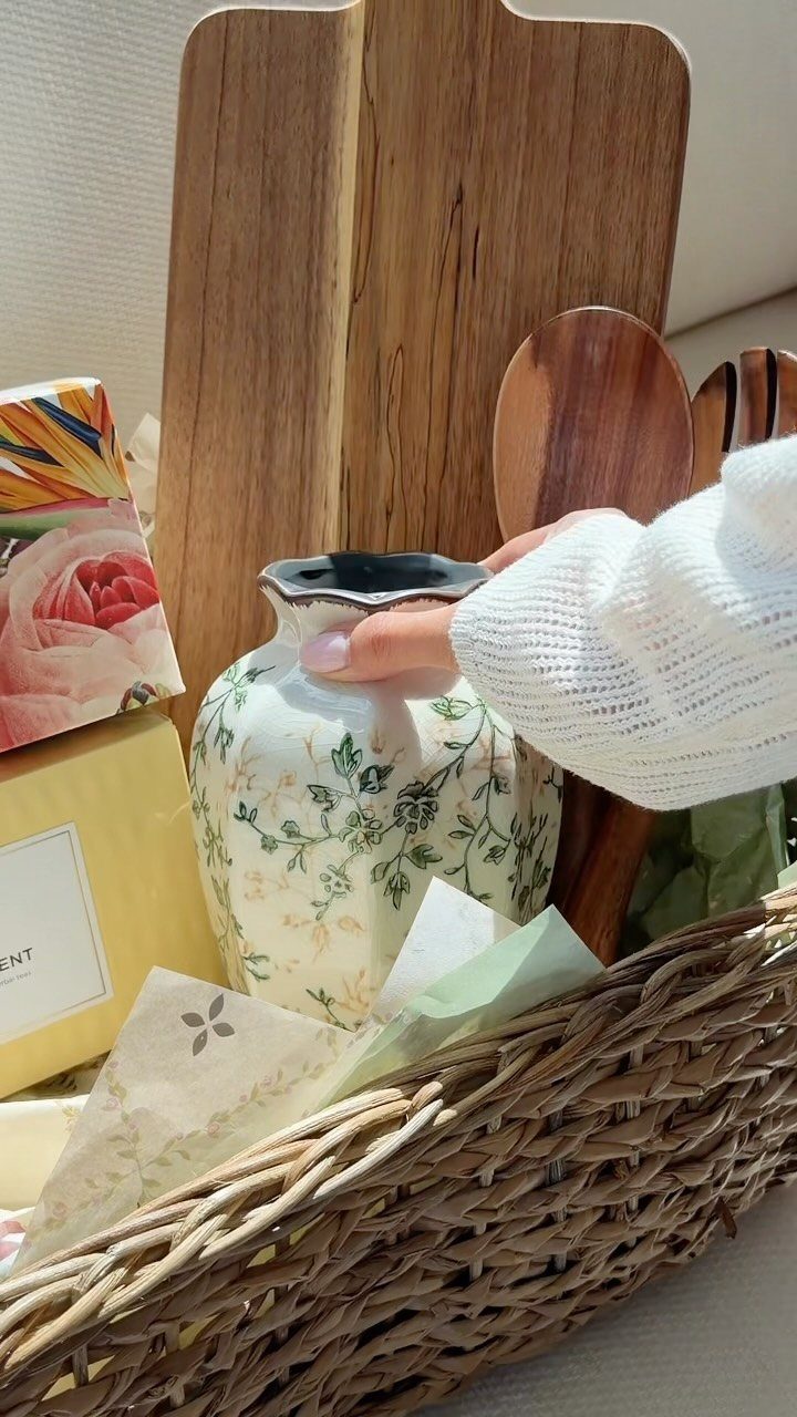 Woven gift basket containing a floral pitcher, wooden board, and wrapped presents.