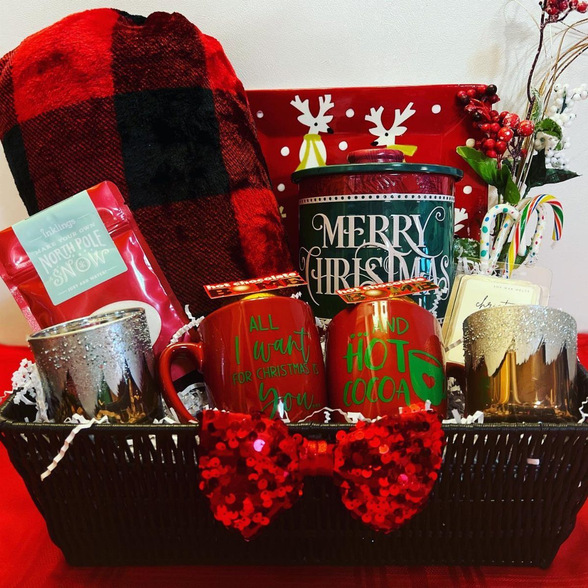 Christmas gift basket with red hot cocoa mugs, plaid blanket, and festive treats.