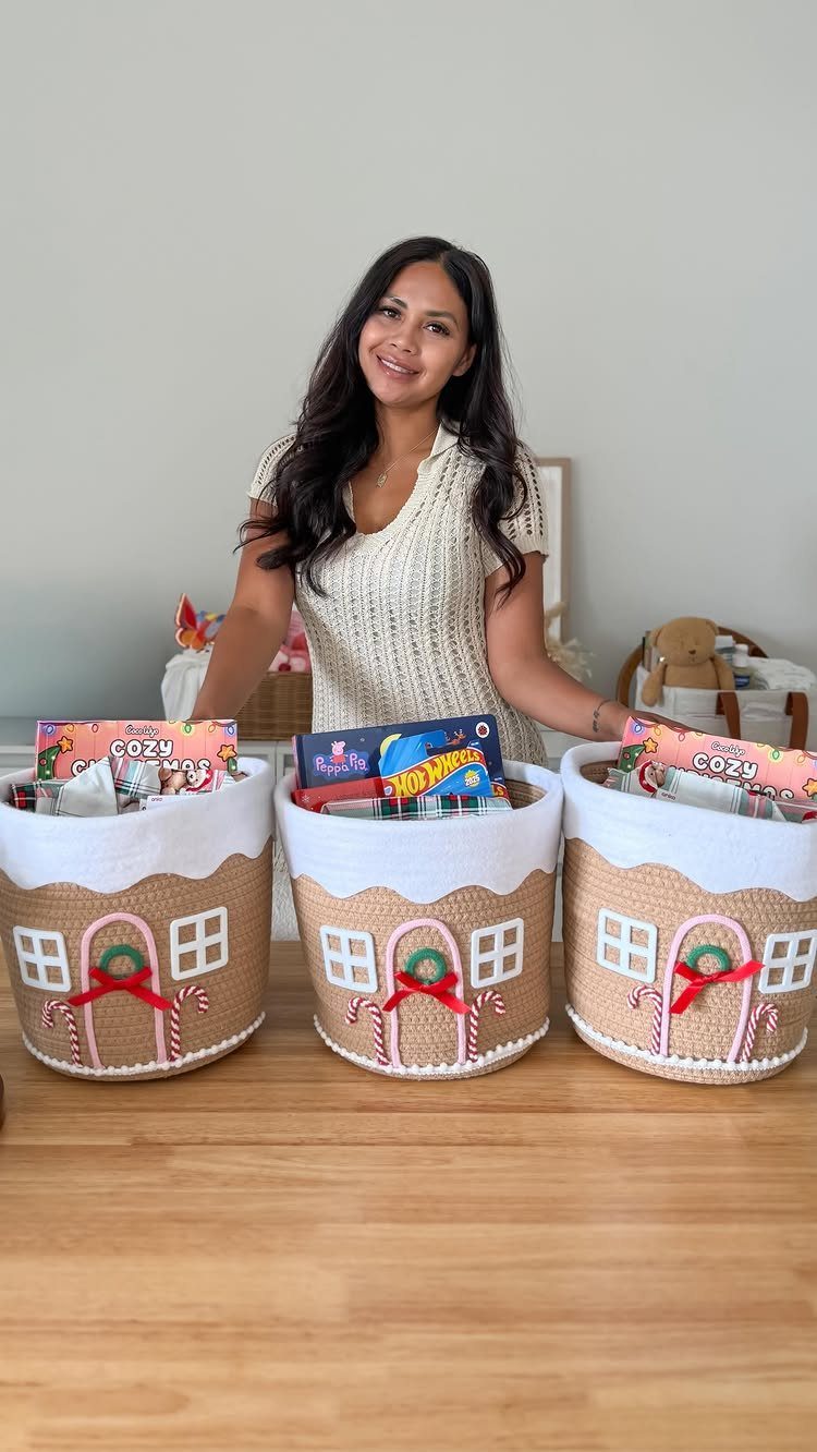 Woman standing behind three gingerbread house themed Christmas Eve baskets filled with gifts.