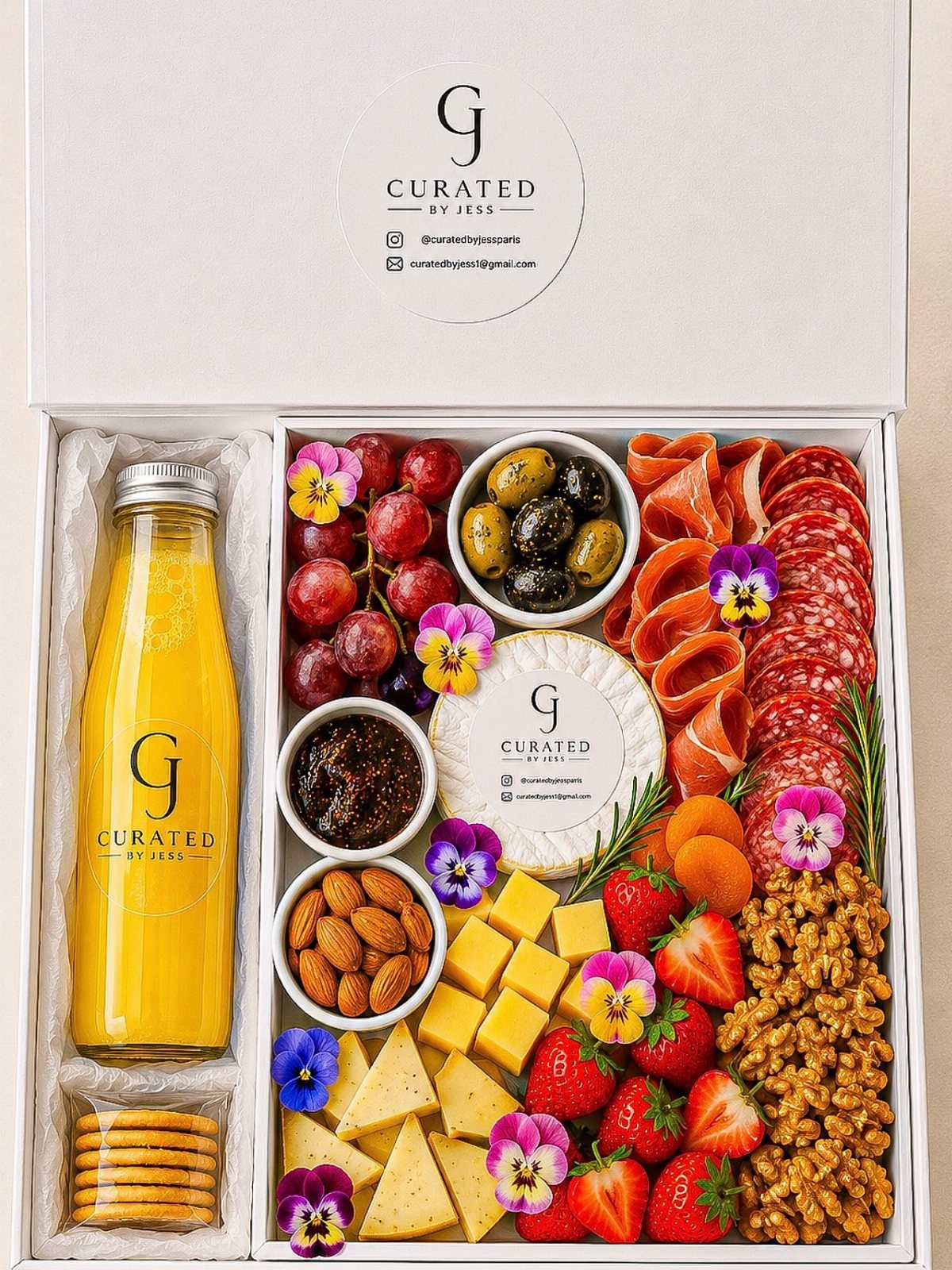 Gourmet meat and cheese sampler box with fruit, nuts, and a juice bottle.