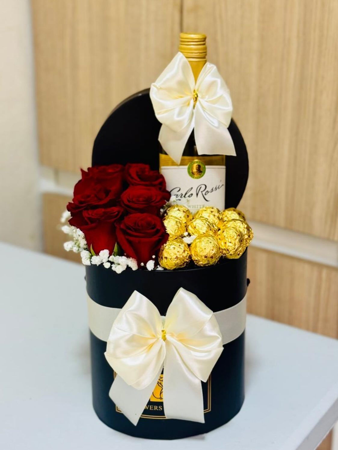 Wine bottle, red roses, and gold chocolates in a black gift box.