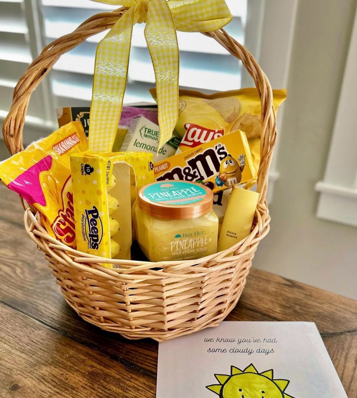 Wicker gift basket filled with yellow snacks and a pineapple jar on wood.
