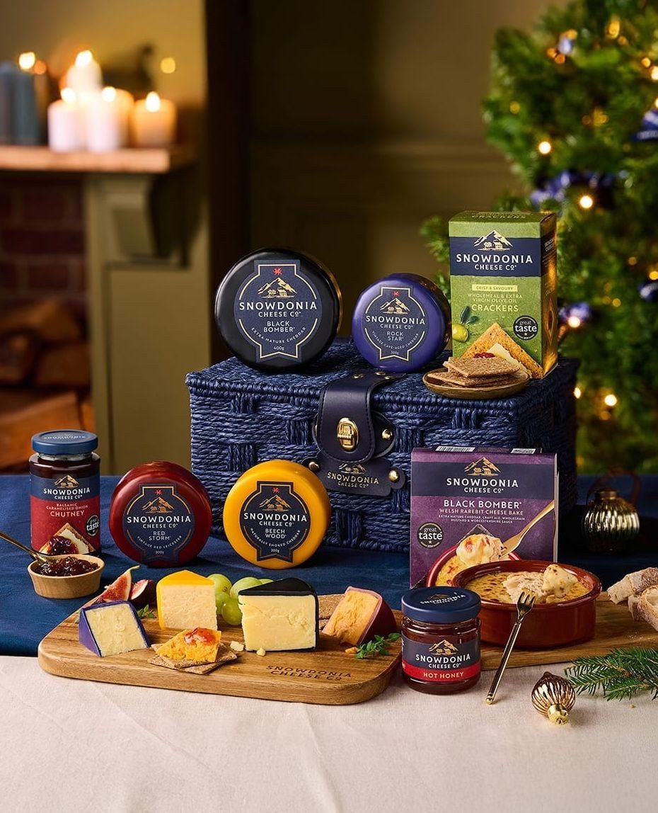 Gourmet cheese gift hamper with various cheeses, crackers, and chutneys near a Christmas tree.