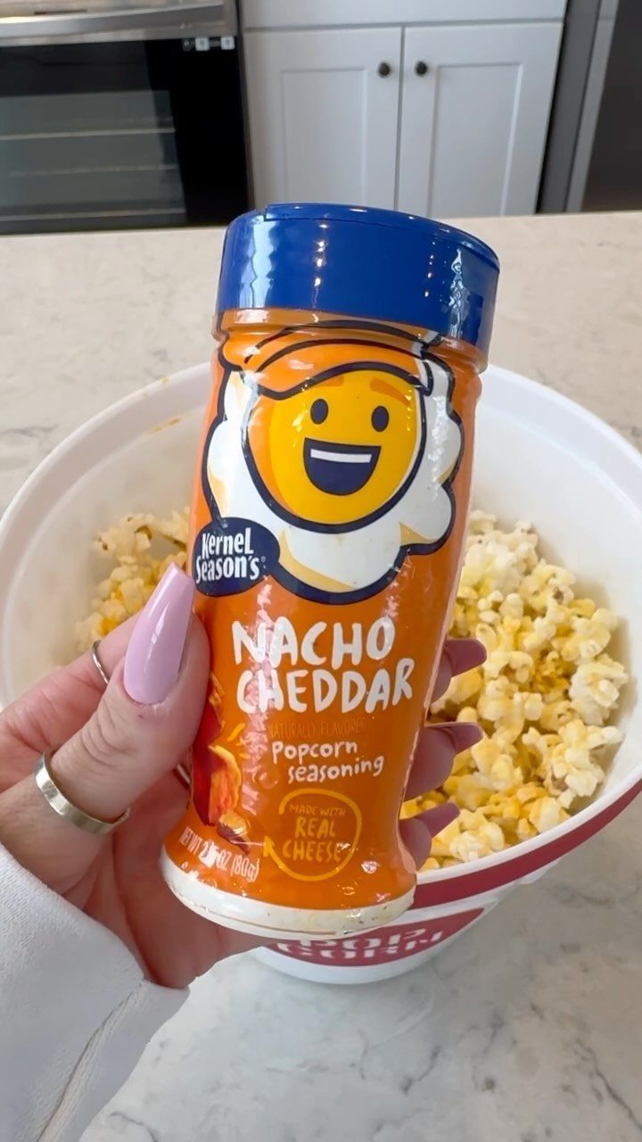 Nacho Cheddar popcorn seasoning bottle held in front of a bowl of popcorn.