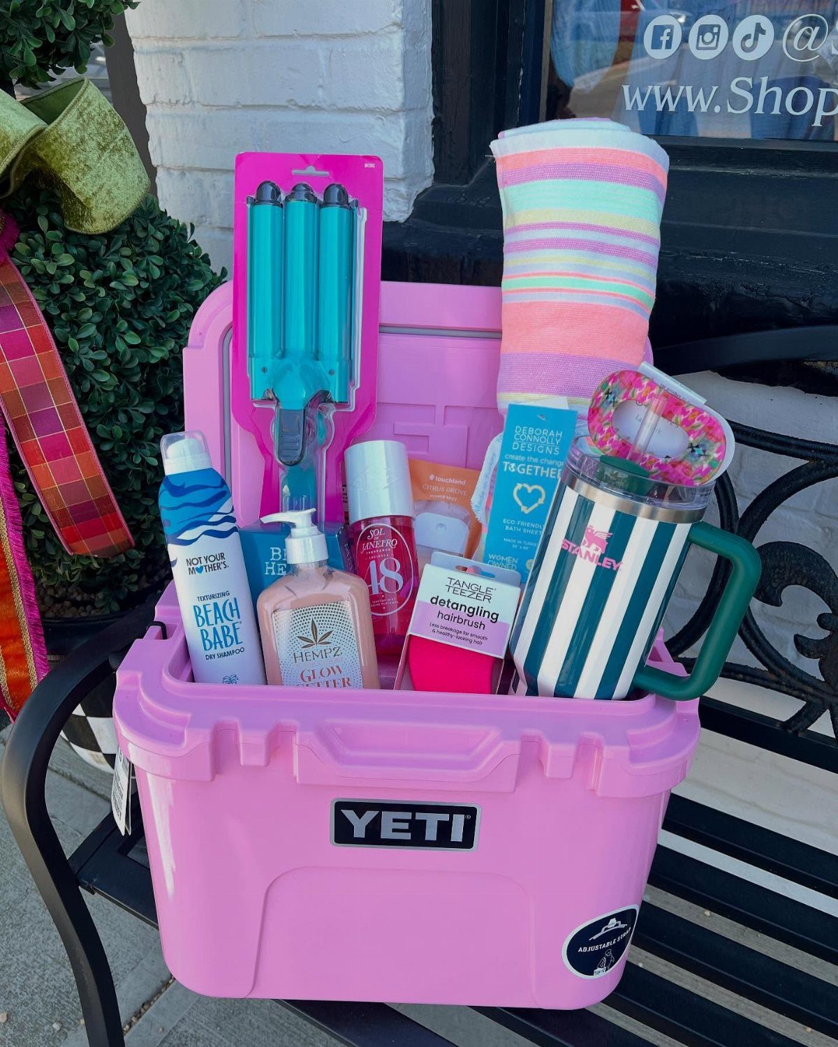 Pink Yeti cooler filled with beach towels, sunscreen, and a striped tumbler on a bench.
