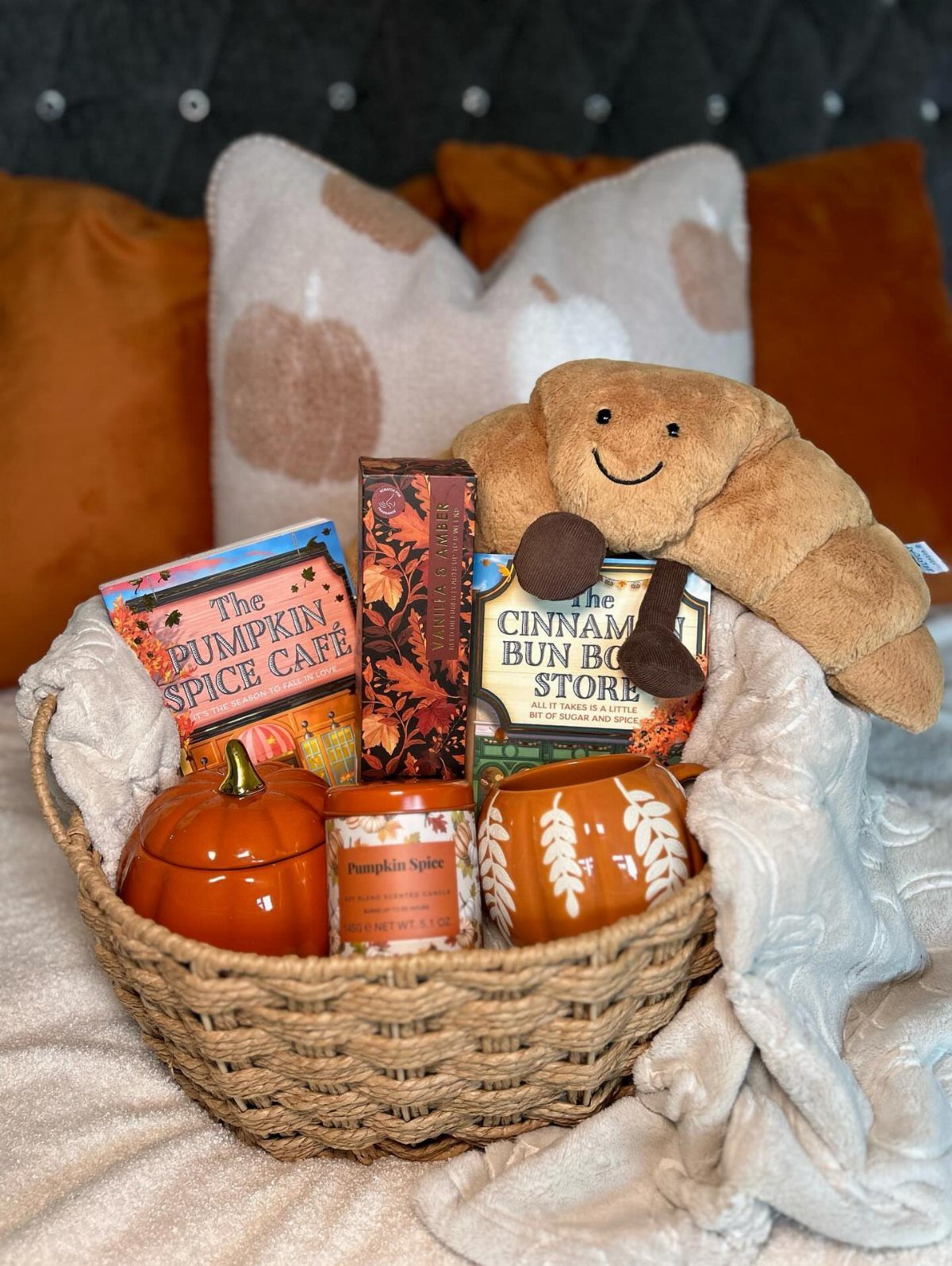 Woven gift basket containing autumn books, a candle, and a plush croissant toy.