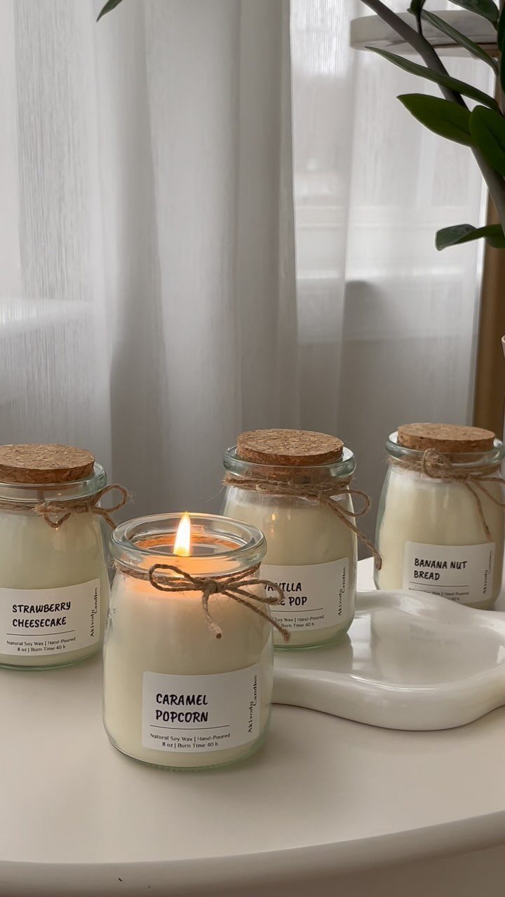 Four dessert-scented candles in glass jars with cork lids on a white table.