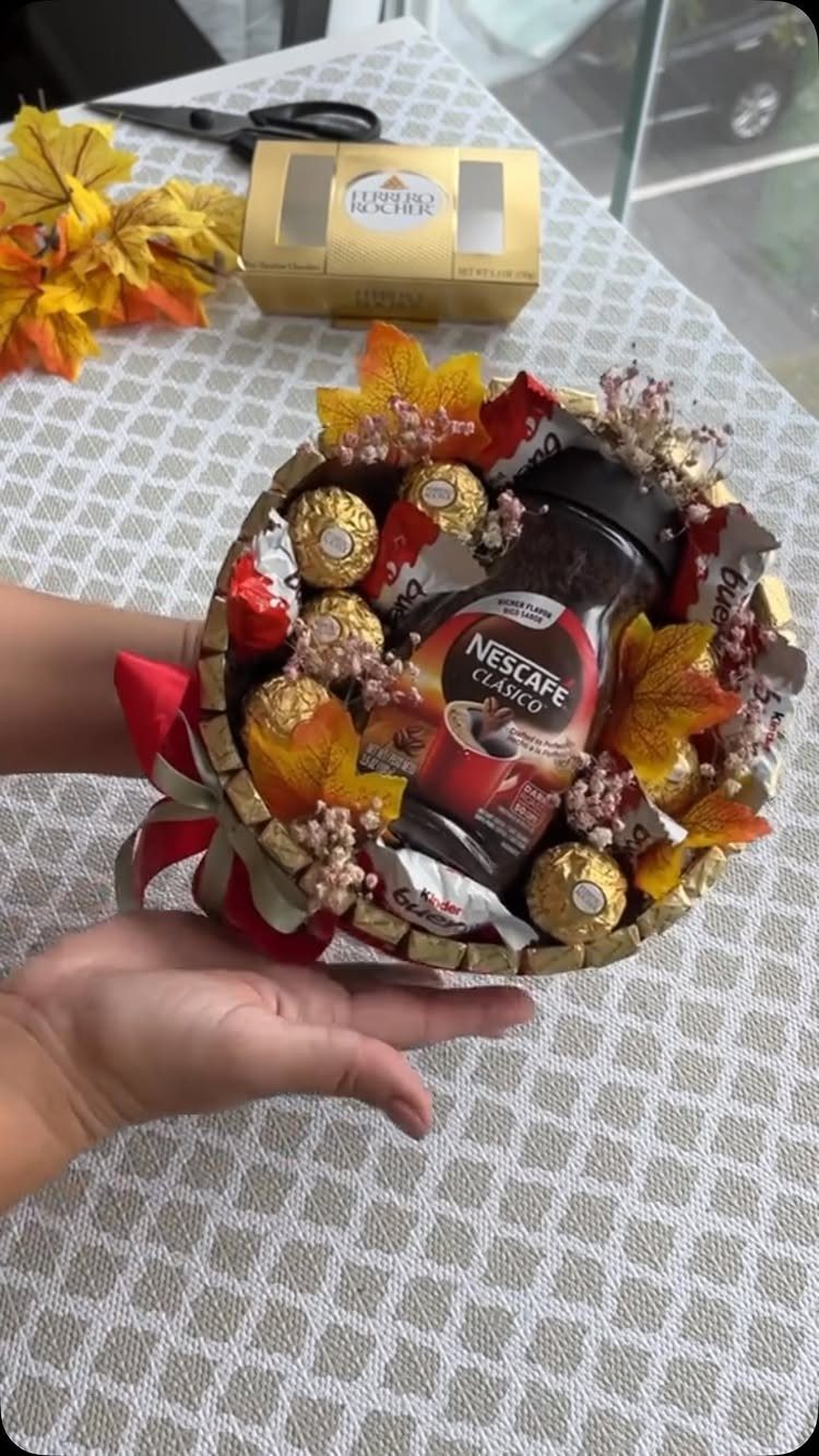 Small gold-rimmed gift basket containing coffee, chocolates, and autumn leaves held by hands.