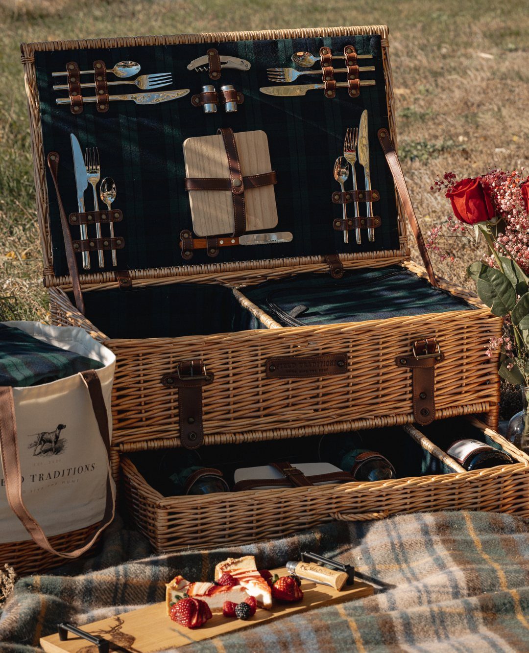 Wicker picnic hamper open on a plaid blanket with charcuterie and red roses.