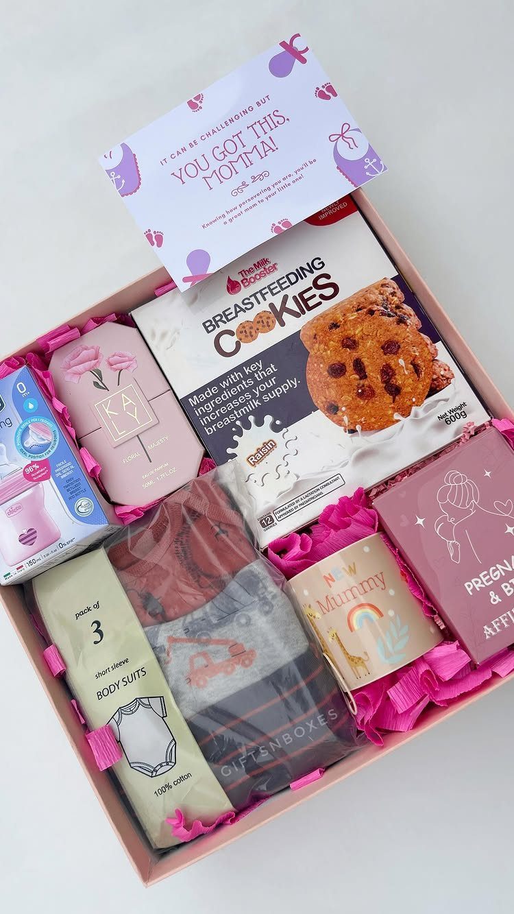 Pink gift box containing breastfeeding cookies, baby clothes, a mug, and a card.
