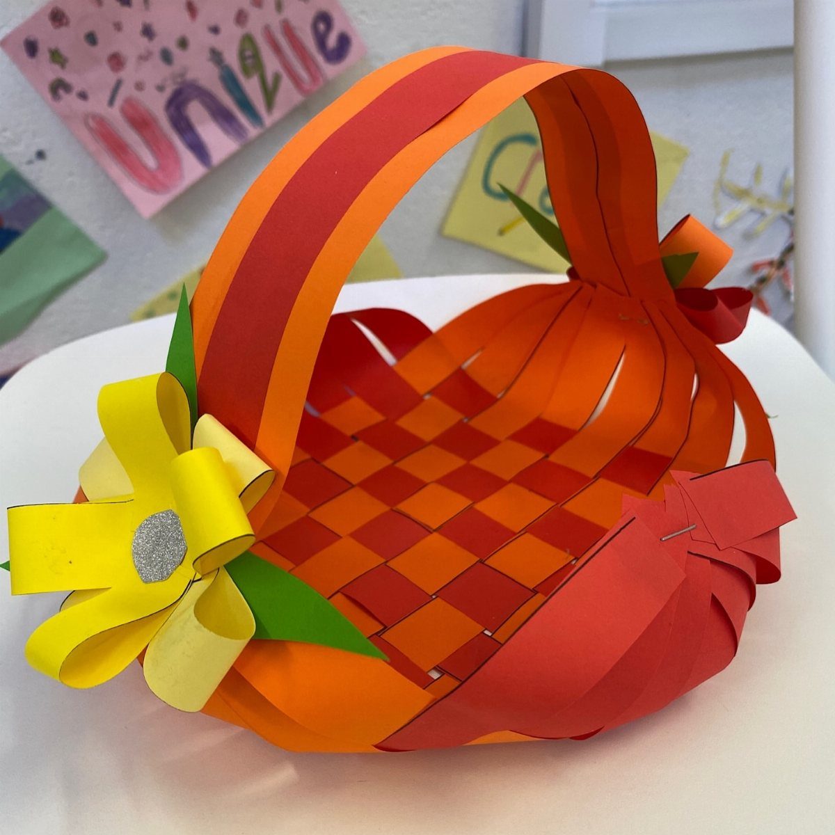 Woven orange and red paper Easter basket with a yellow paper flower decoration.