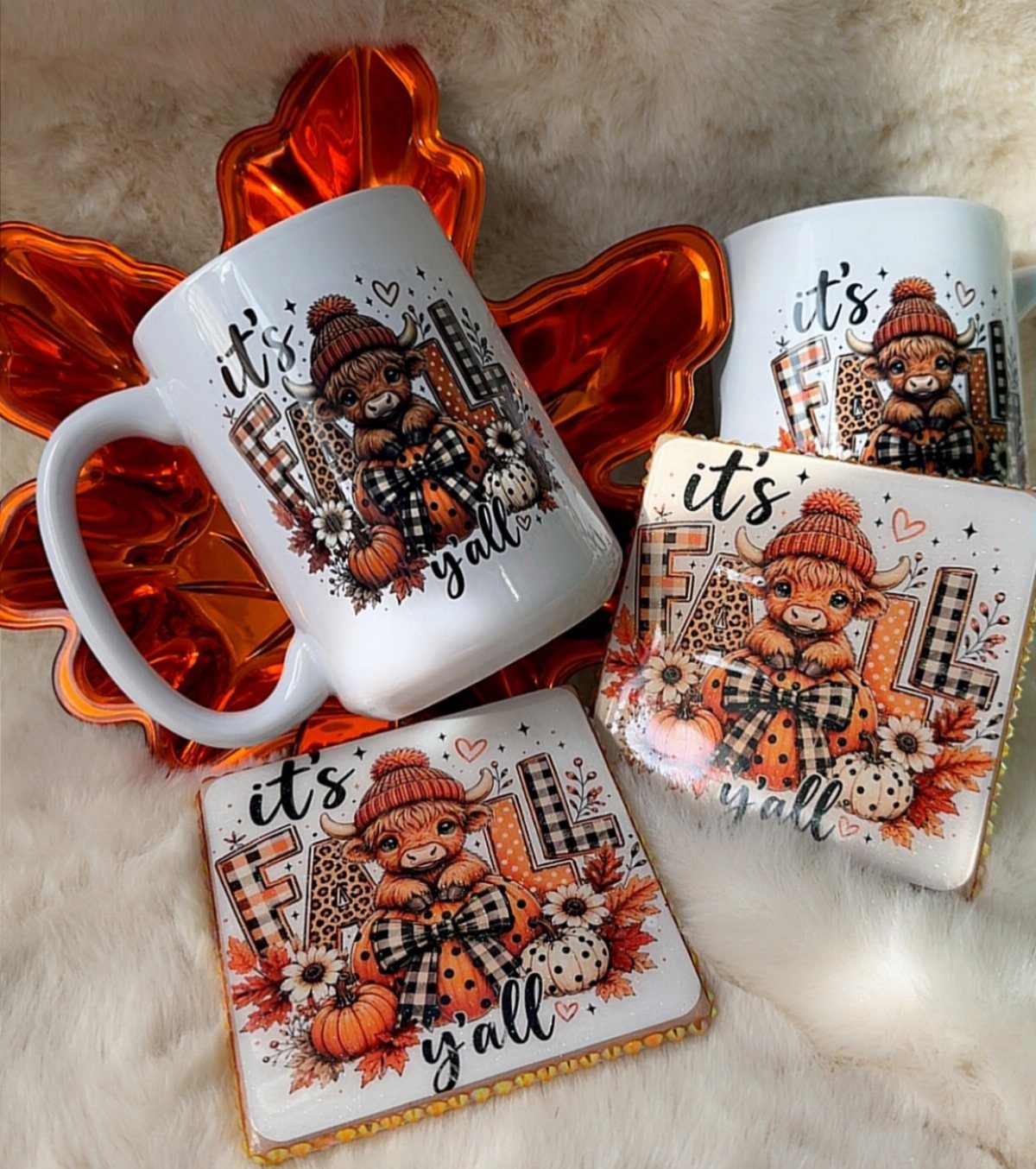 Two white mugs and square coasters with autumn highland cow illustrations on white fur.