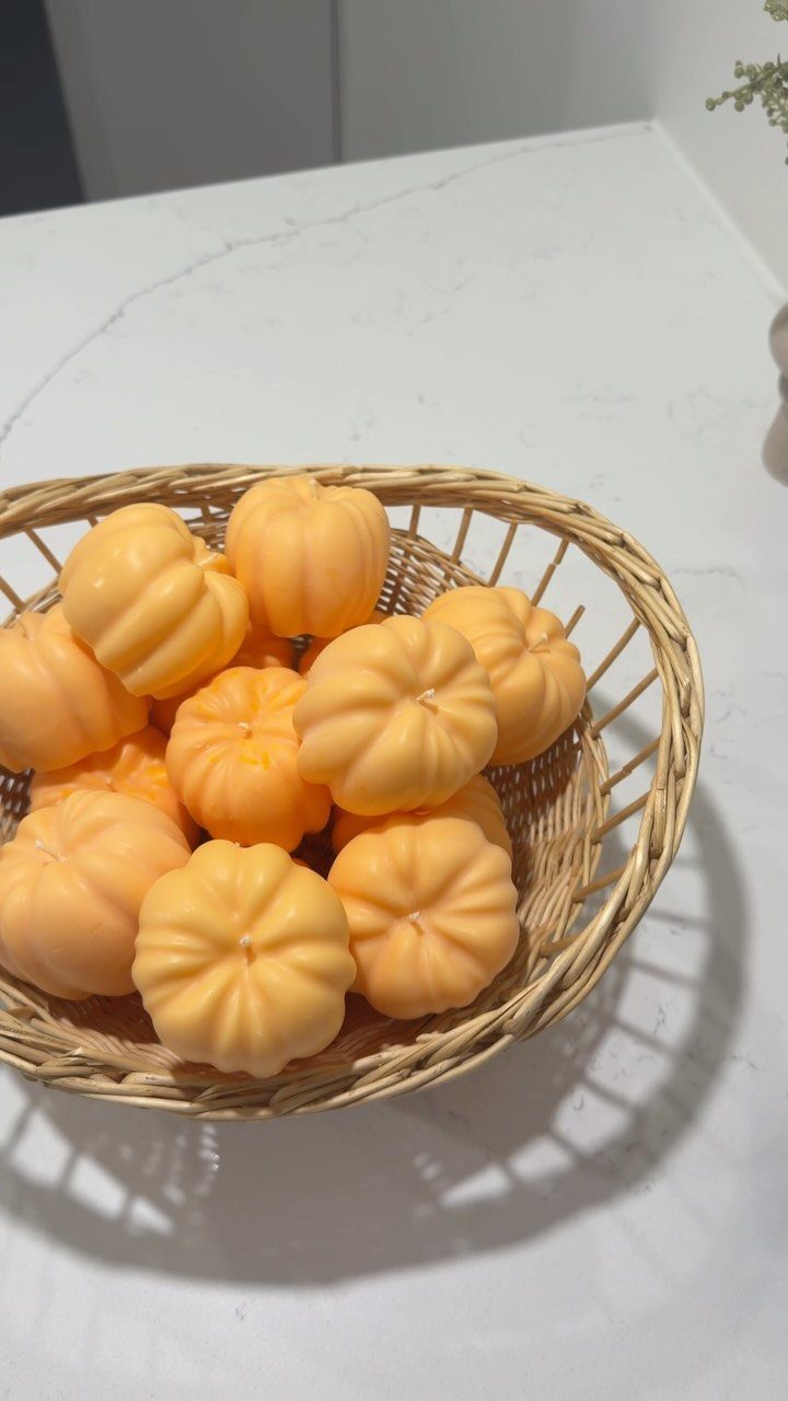Pumpkin-shaped orange candles filling a small woven wicker basket on white marble.
