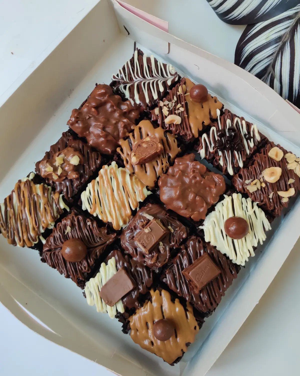 Assorted chocolate brownies with various toppings and drizzles in a white box.
