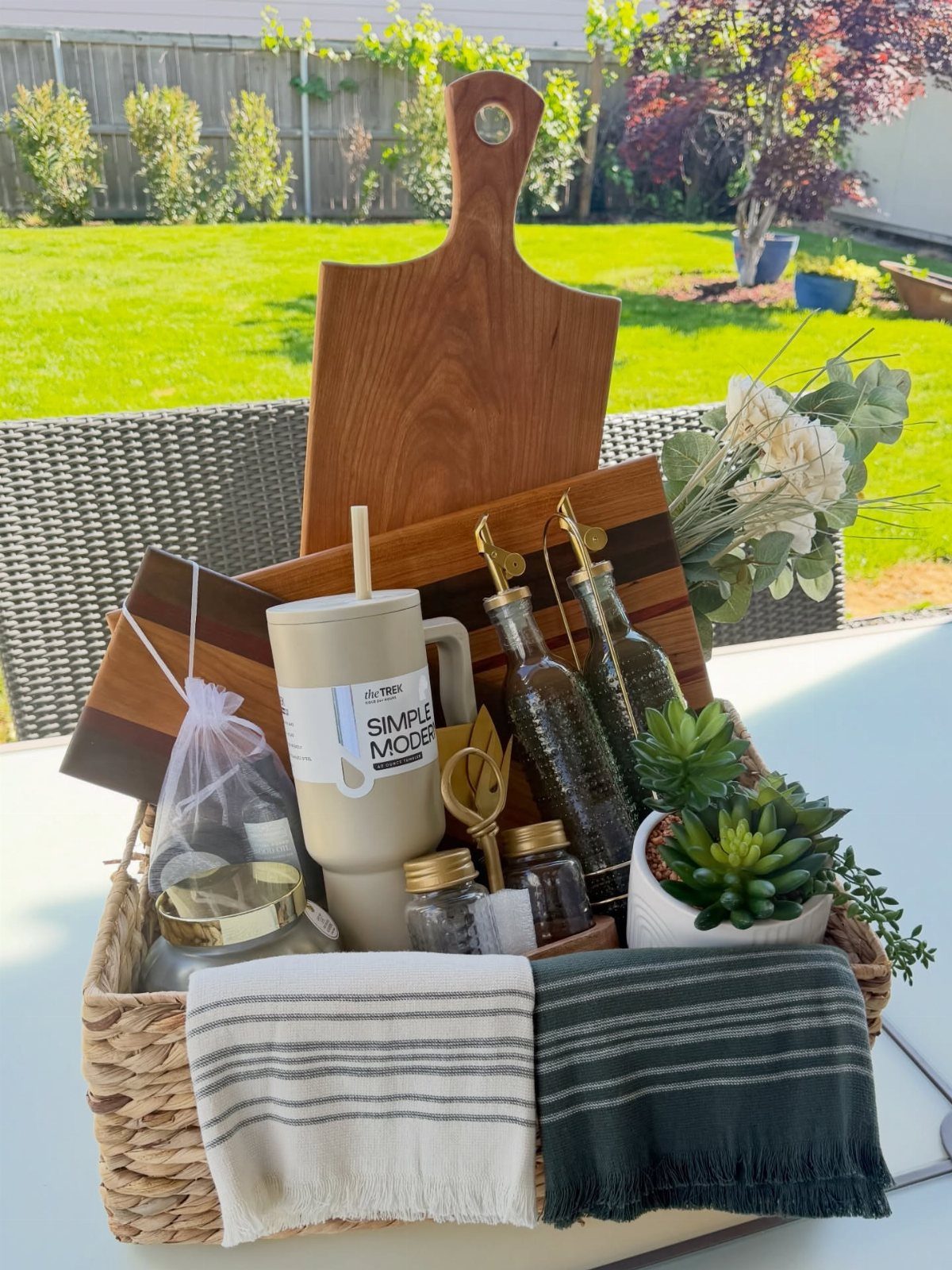 Woven gift basket with wooden cutting board, travel mug, and potted succulent outdoors.
