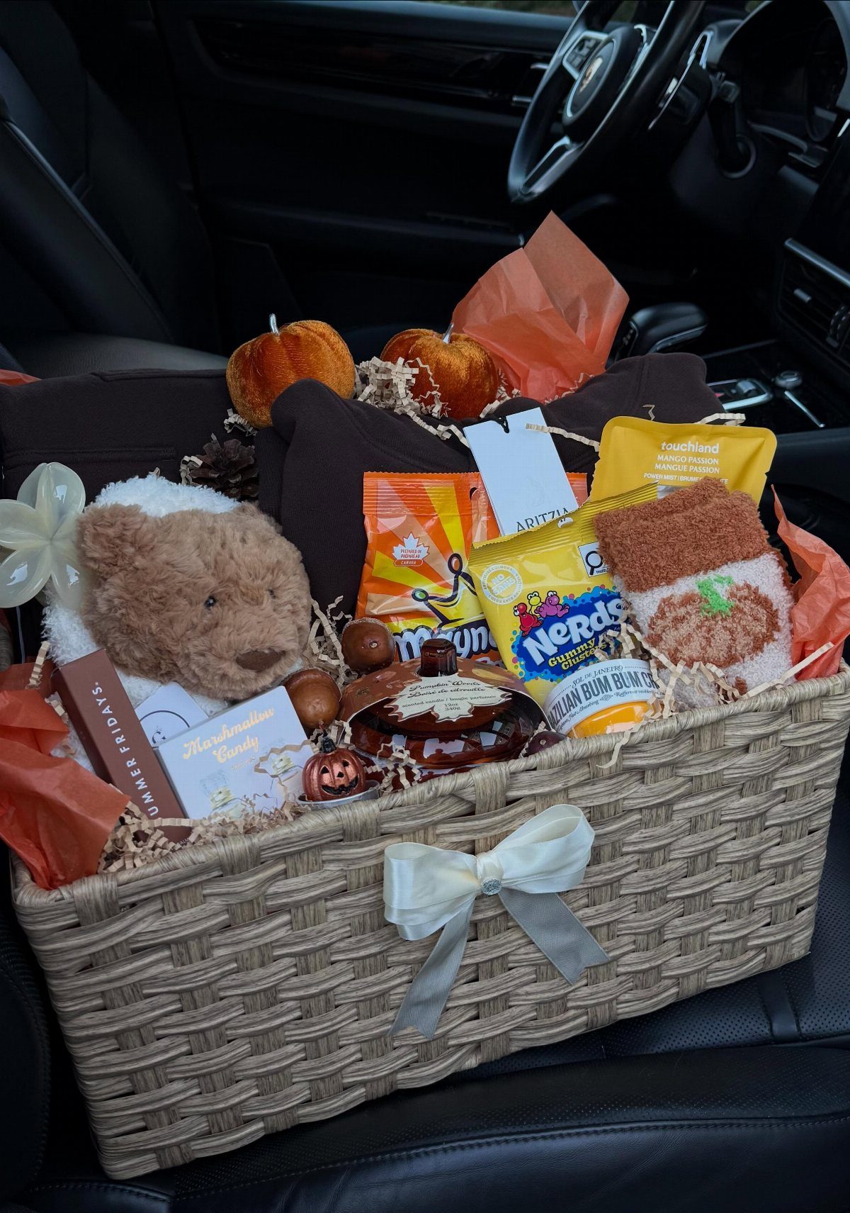 Woven gift basket with snacks, teddy bear, and pumpkins on a car seat.
