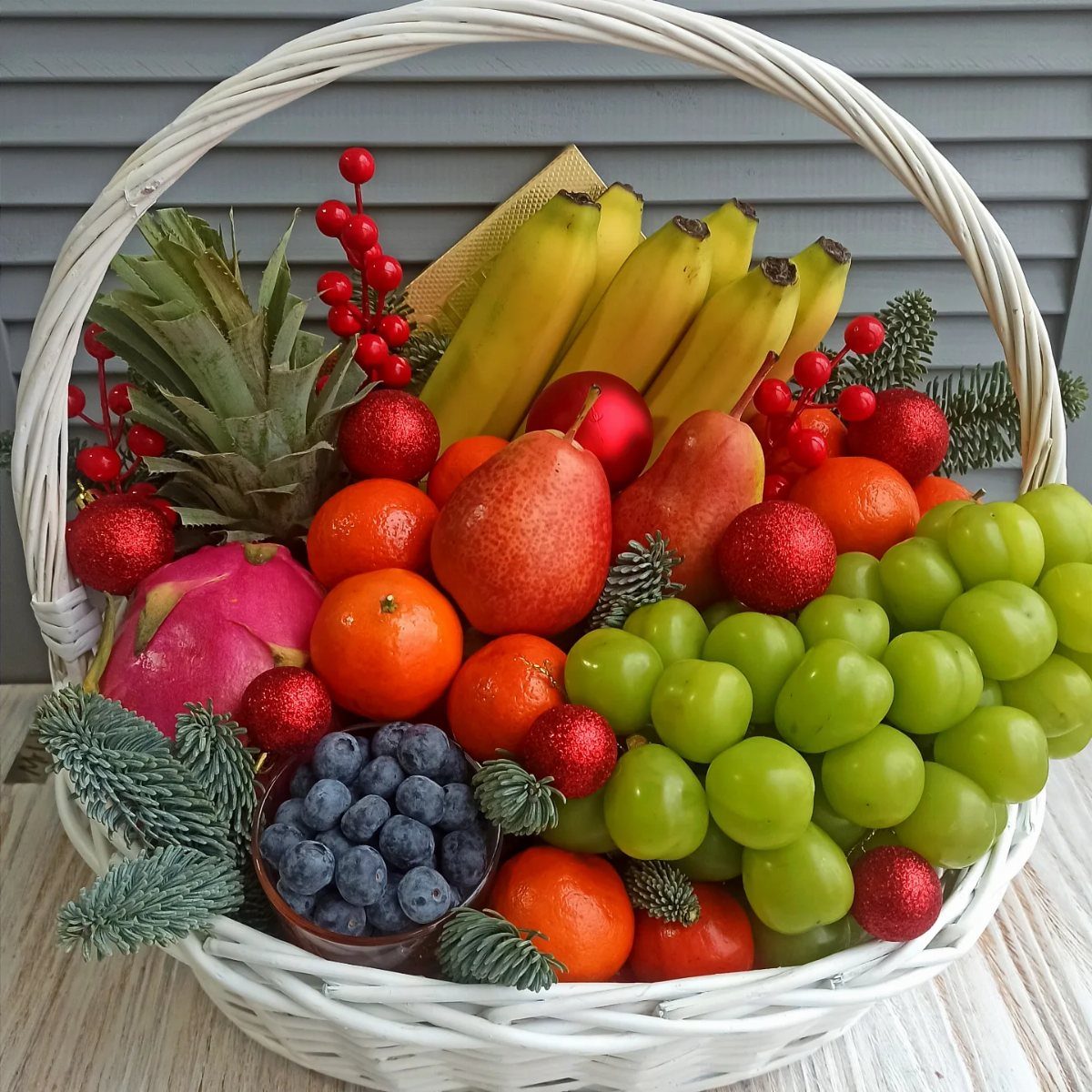 White wicker gift basket filled with assorted fresh fruits and red Christmas ornaments.