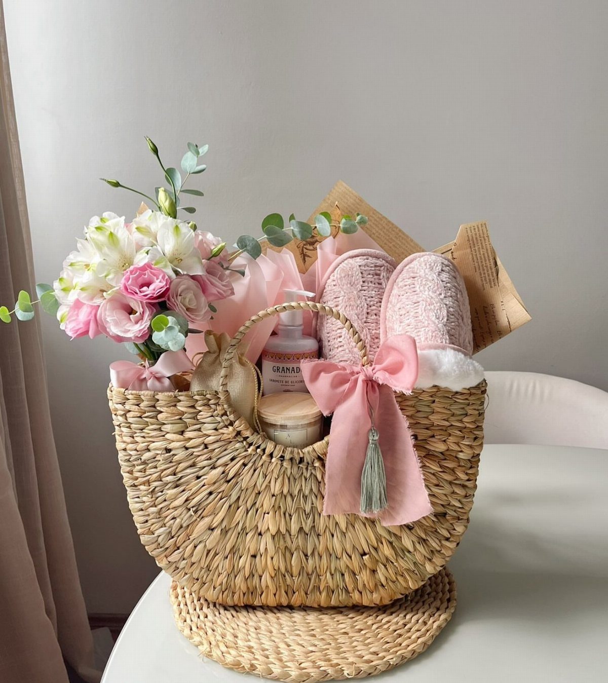 Woven gift basket containing pink slippers, skincare products, and fresh pink flowers.