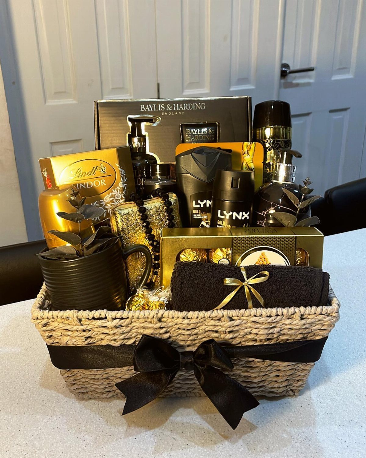Woven gift basket filled with black and gold toiletries, chocolates, and a mug.