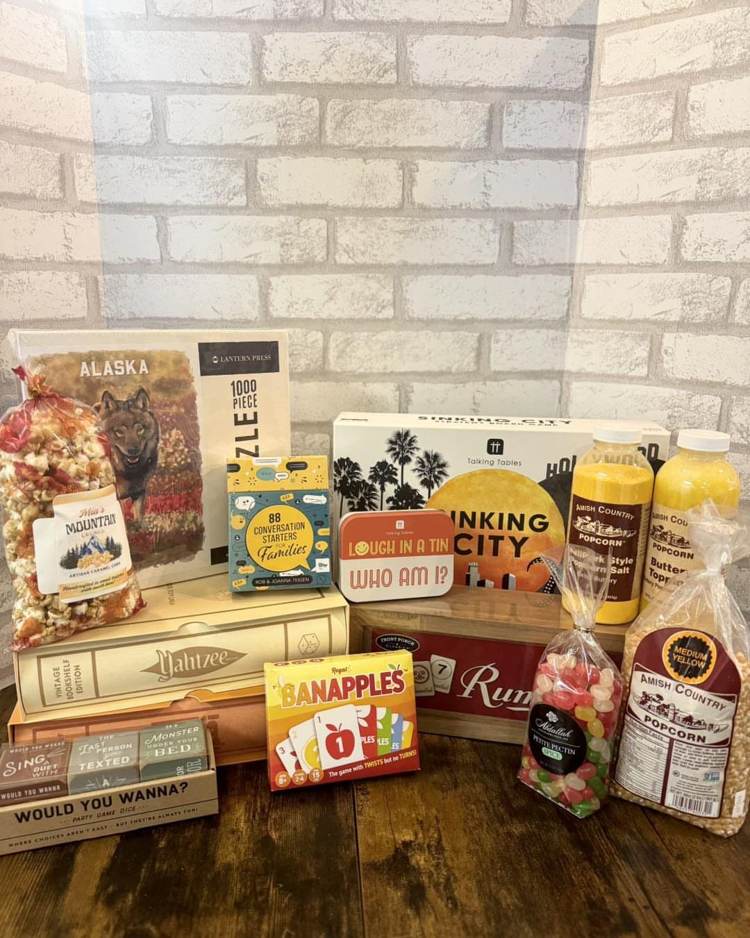 Gift basket items including a puzzle, card games, popcorn, and drinks on wood.
