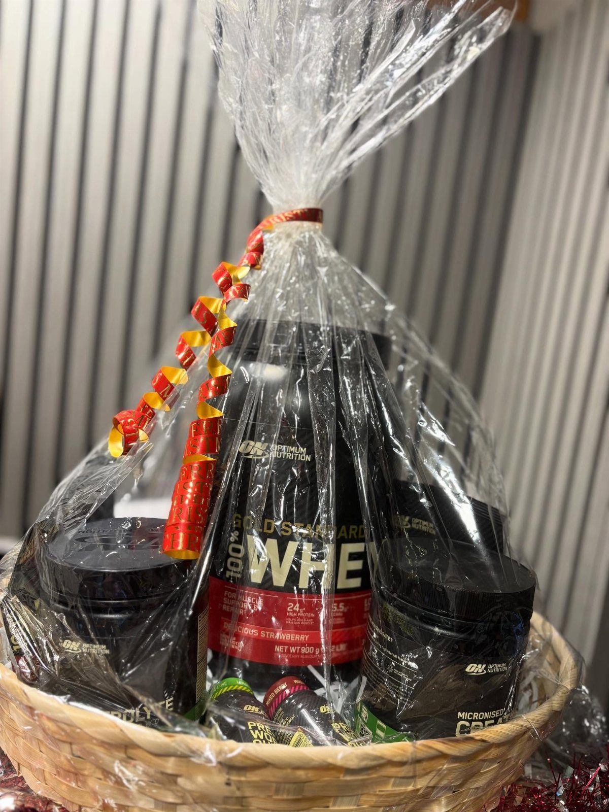 Wicker gift basket wrapped in cellophane containing whey protein and fitness supplements.