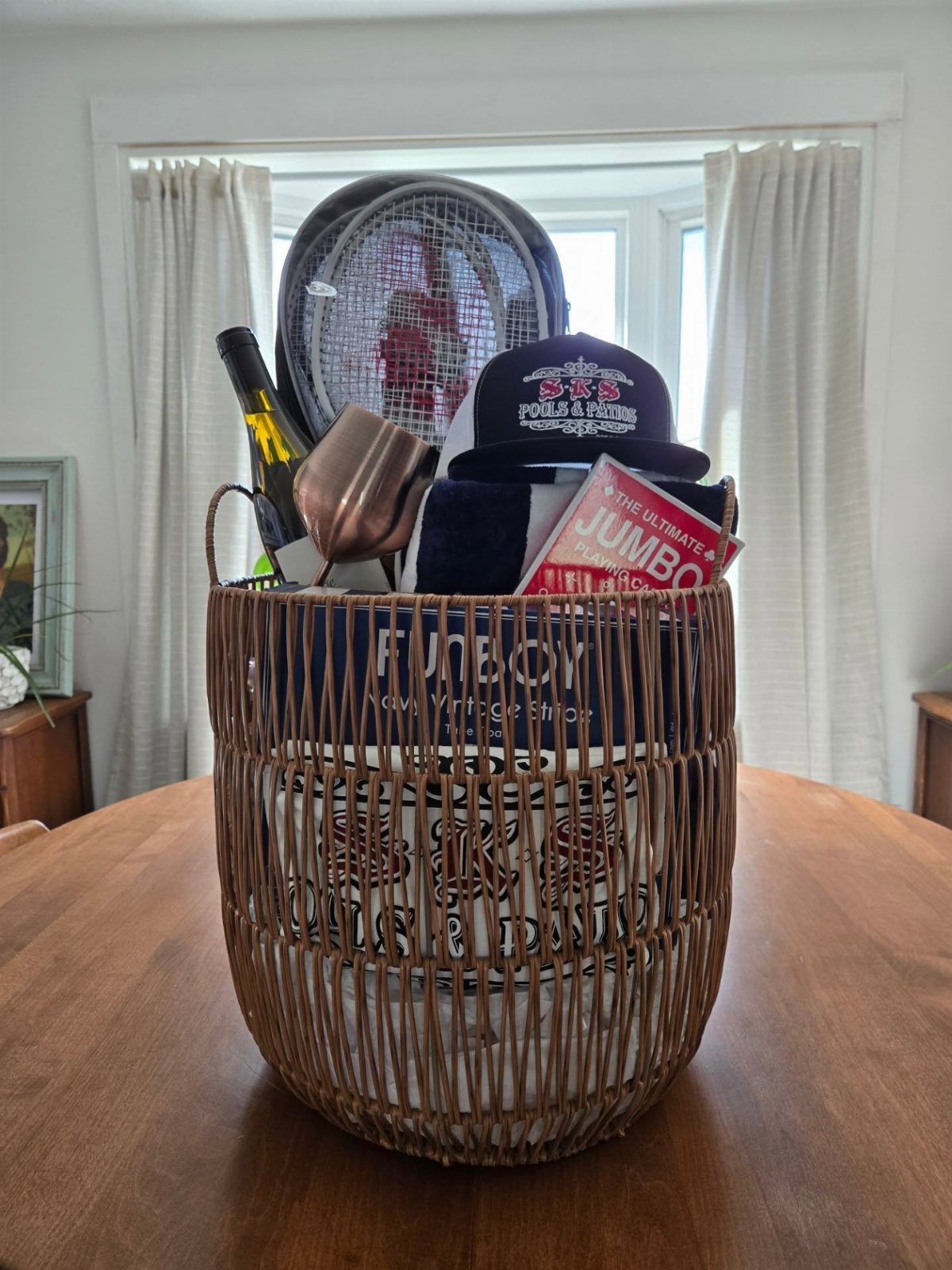 Woven gift basket filled with a tennis racket, baseball cap, and wine bottle.