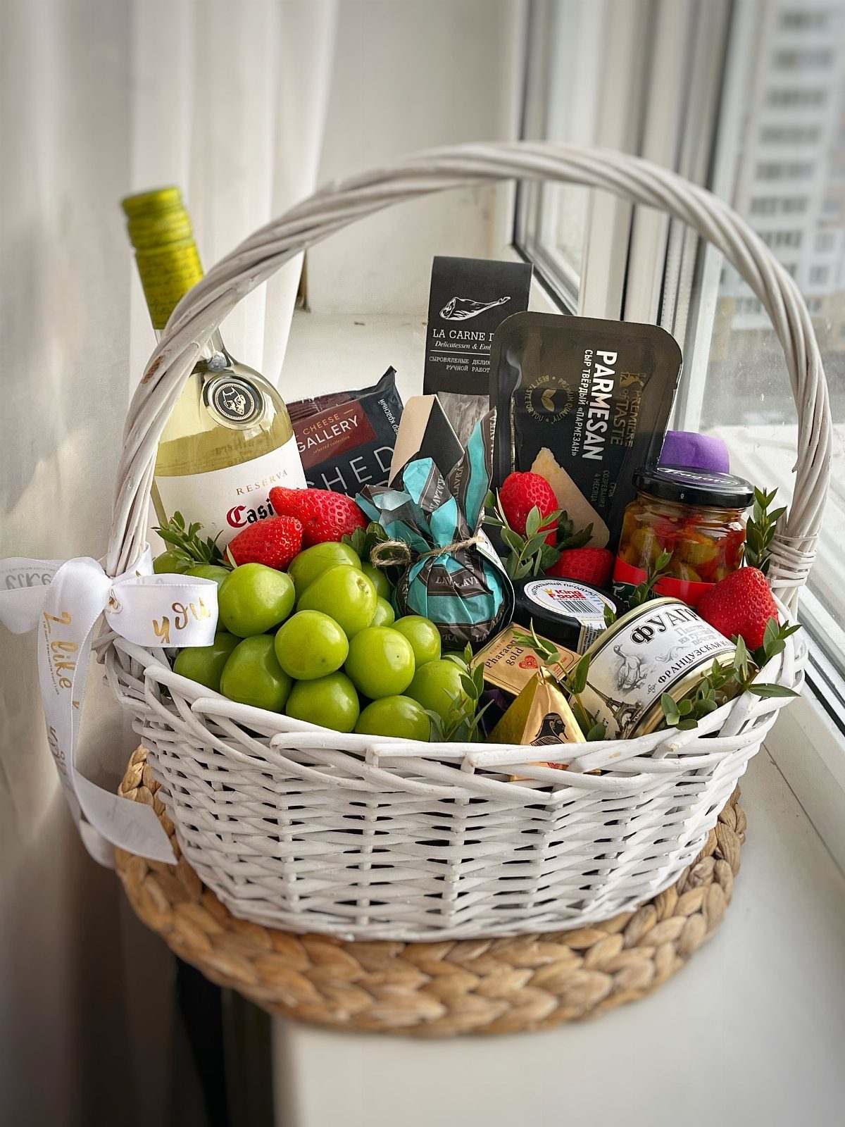White wicker gift basket containing white wine, assorted cheeses, green grapes, and strawberries.